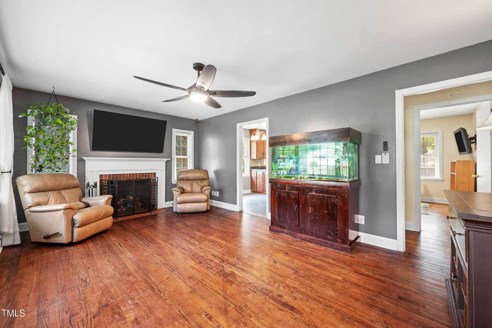 A spacious living room with wood floors, gray walls, two tan recliners by a fireplace, a large wall-mounted TV, a tall aquarium, and large windows with white curtains letting in natural light.