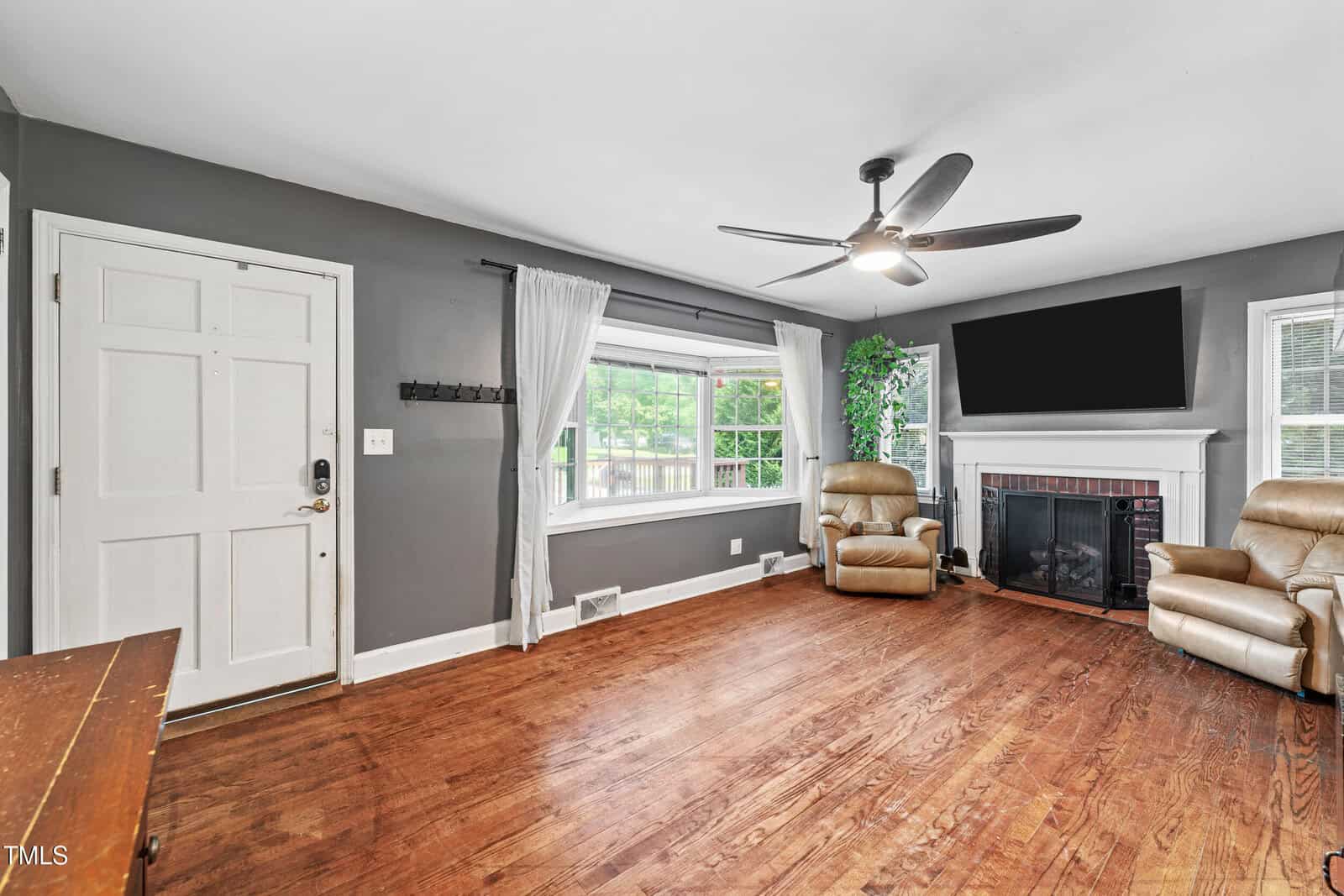 A spacious living room with hardwood floors, gray walls, a ceiling fan, two beige recliners by a fireplace, large windows with white curtains, and a flat-screen TV above the mantel.