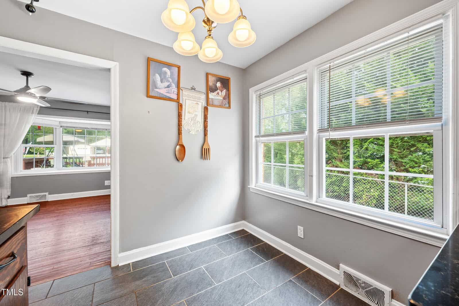 A small dining area with gray walls, a window letting in natural light, dark tile flooring, and decorative wall art. Wooden utensils hang on the wall beside framed pictures. An adjacent room with wood floors is visible.