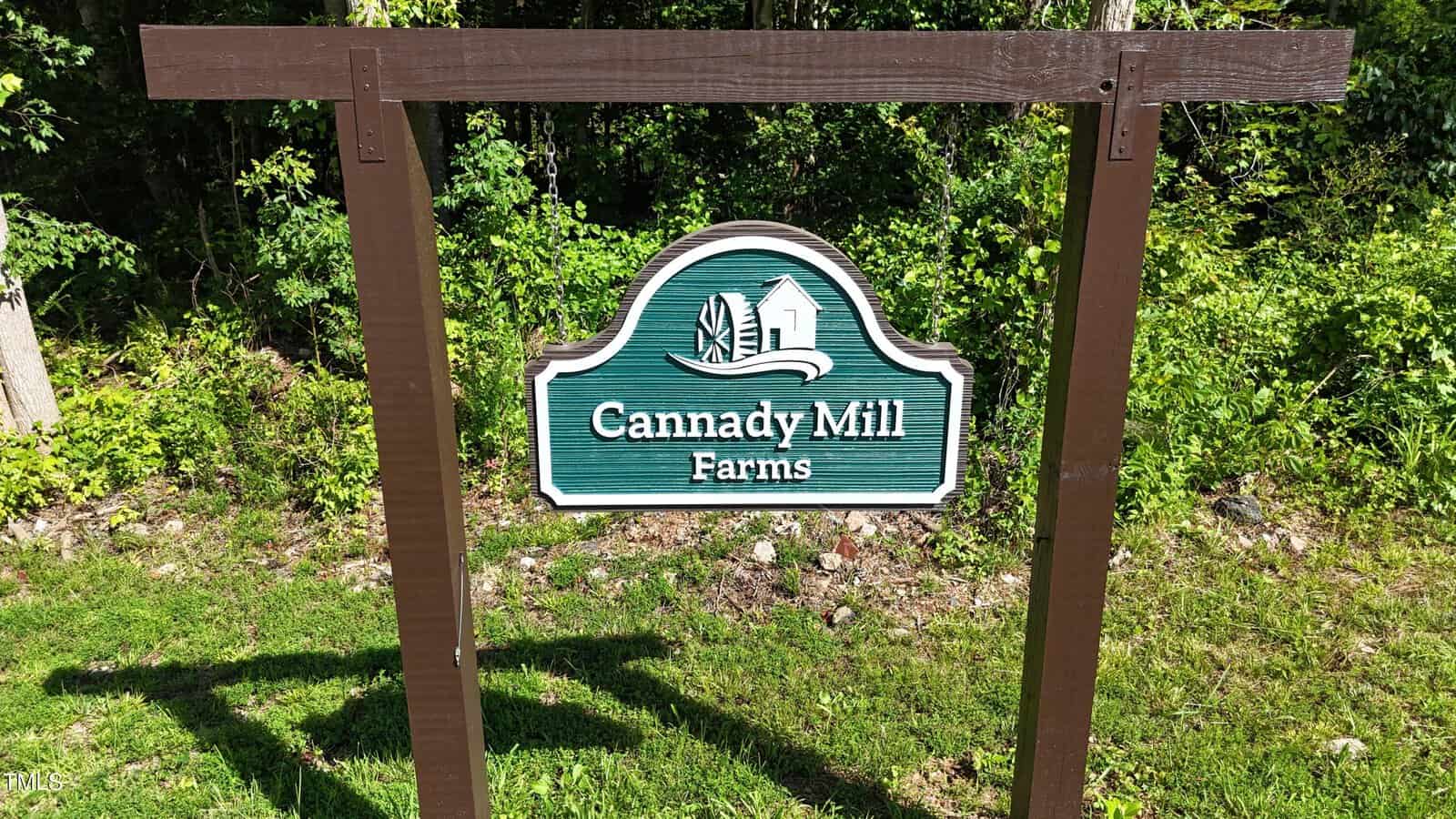 A green sign with white text reads “Cannady Mill Farms” and features a barn and mill illustration, mounted on a wooden frame in front of trees and greenery.