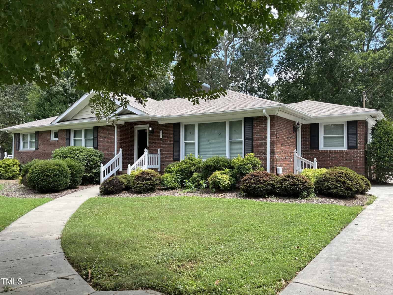 Single-story red brick house with white trim and shutters, a front porch with white railings, and a curved sidewalk. The house is surrounded by green bushes, trees, and a well-kept lawn.