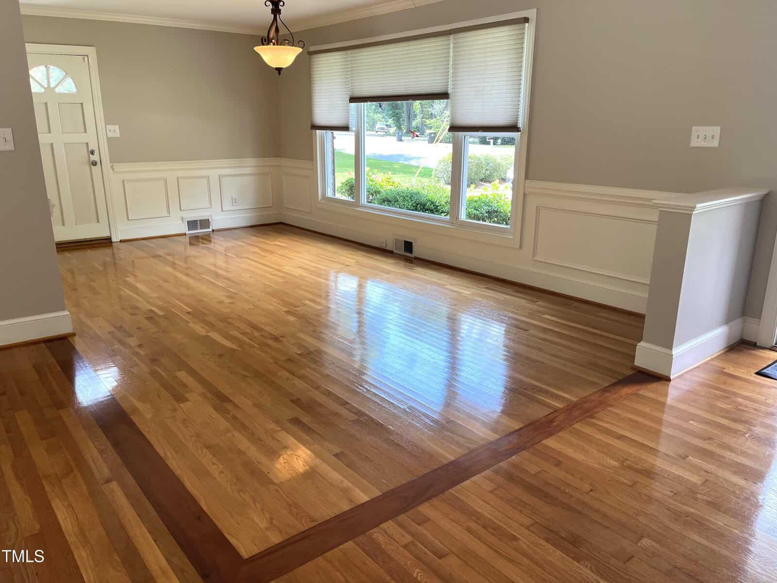 An empty room with light hardwood floors, a darker wood border, large front windows, white trim, and a hanging light fixture. The front door is visible on the left, and sunlight brightens the space.