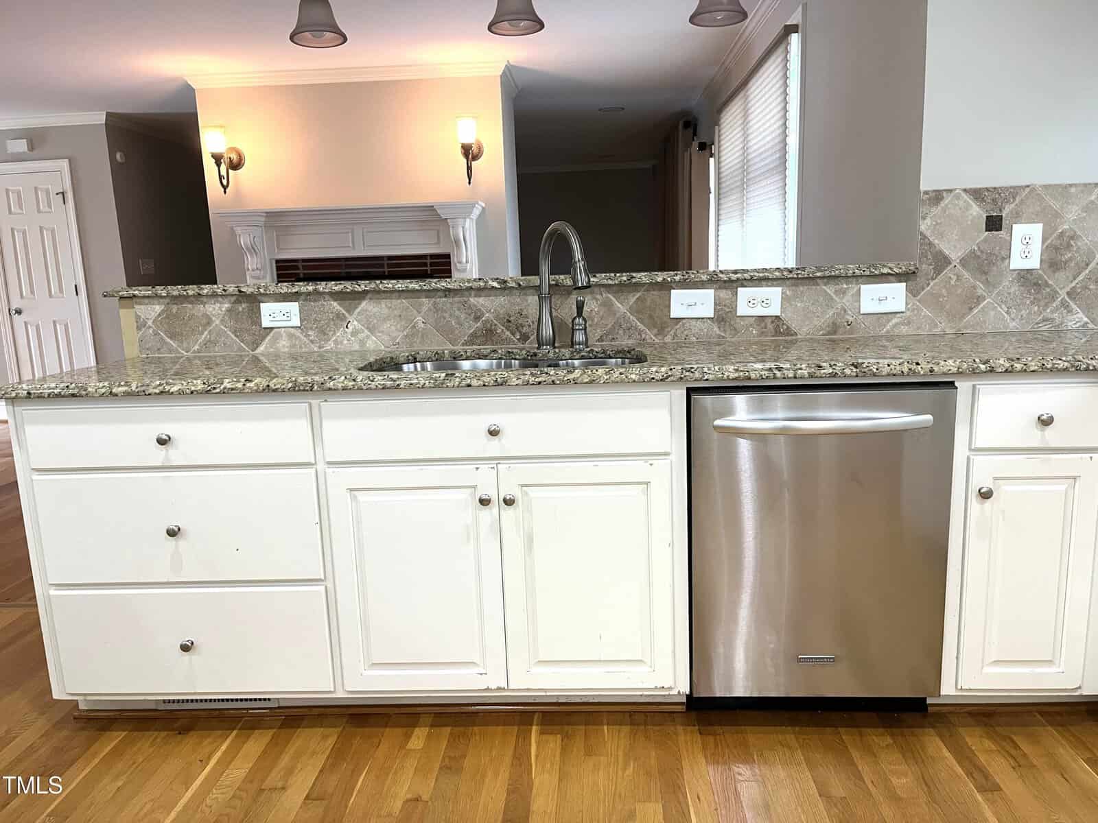 A kitchen island with white cabinets, a stainless steel dishwasher, granite countertop, tile backsplash, and a double sink with a faucet; hardwood floor and wall sconces in the background.