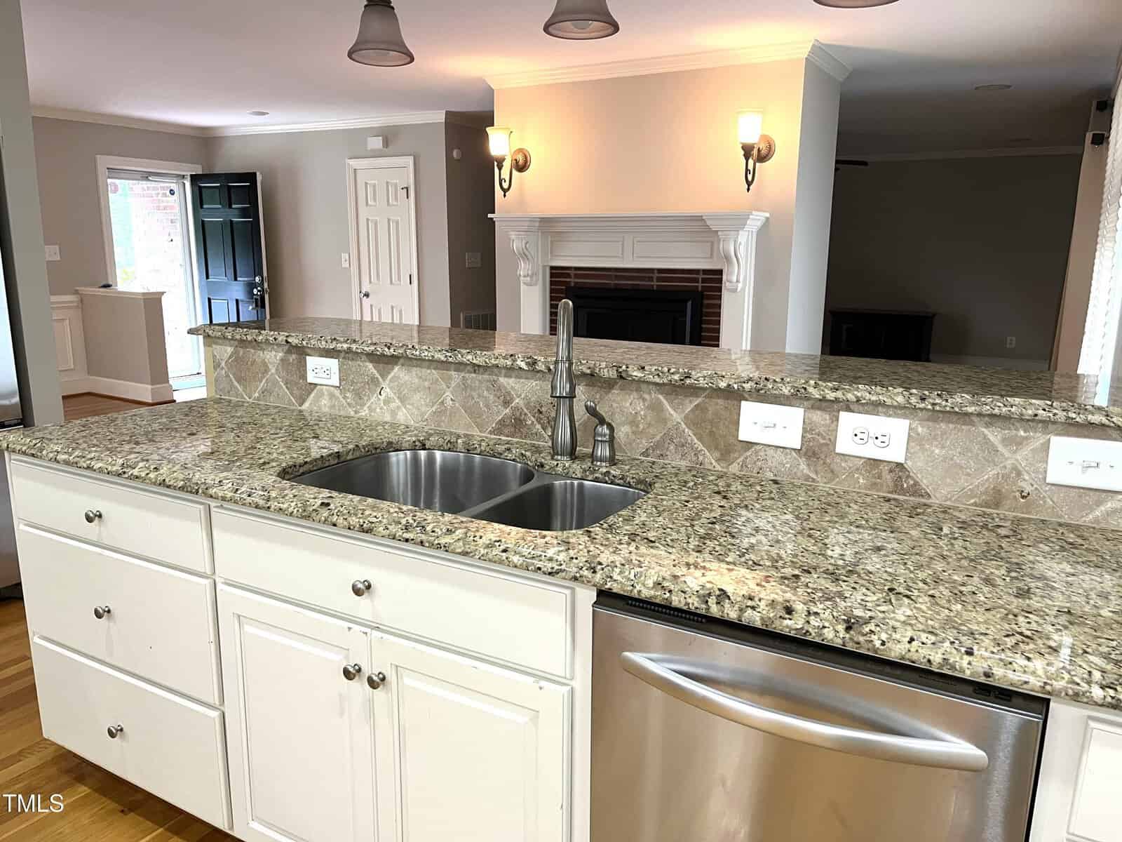 Modern kitchen with granite countertops, double sink, white cabinets, stainless steel dishwasher, and a partial view of a living area with a fireplace and wall sconces in the background.