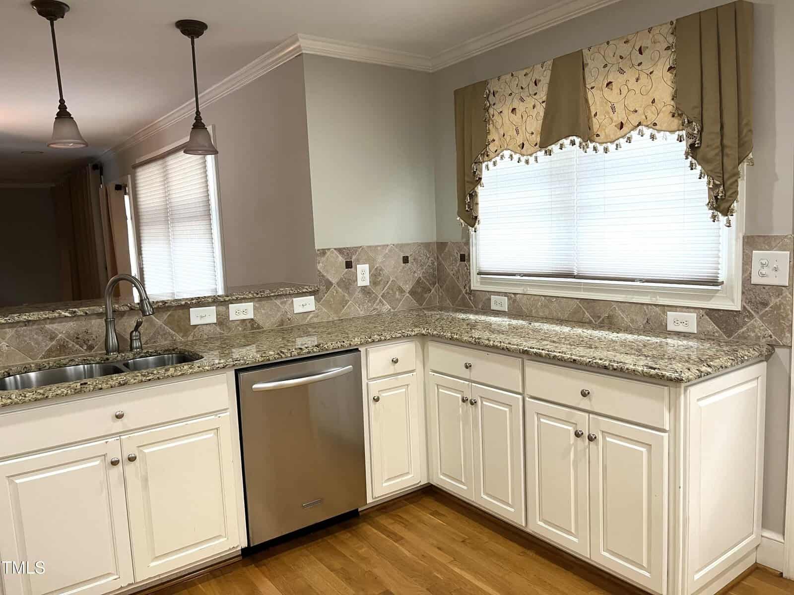 Kitchen with white cabinets, granite countertops, stainless steel dishwasher, double sink, light brown tile backsplash, and a window with a valance above the counter. Hardwood floor and pendant lights are also visible.
