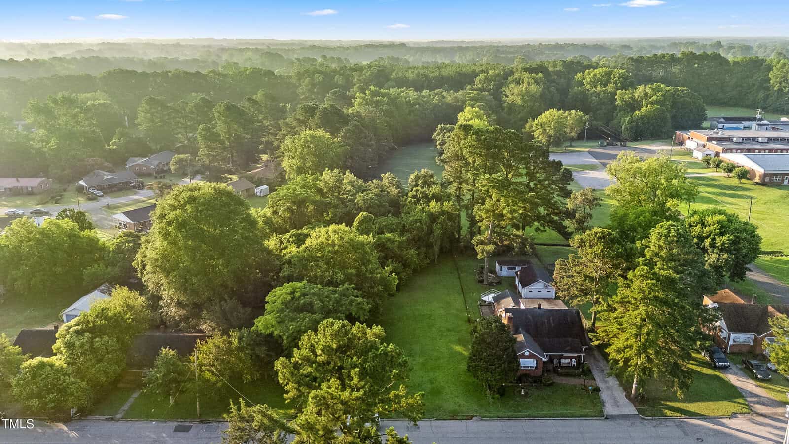 Aerial view of a residential neighborhood with single-family homes, large green lawns, and many tall trees. A school or large building with a field is visible in the upper right. The background shows dense forest.