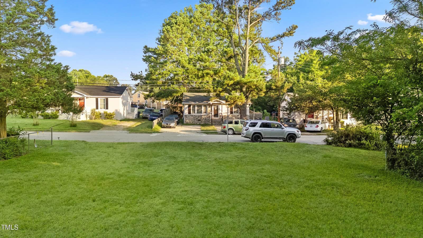A grassy yard with a street and several parked cars in front of single-story houses surrounded by trees under a blue sky with scattered clouds.