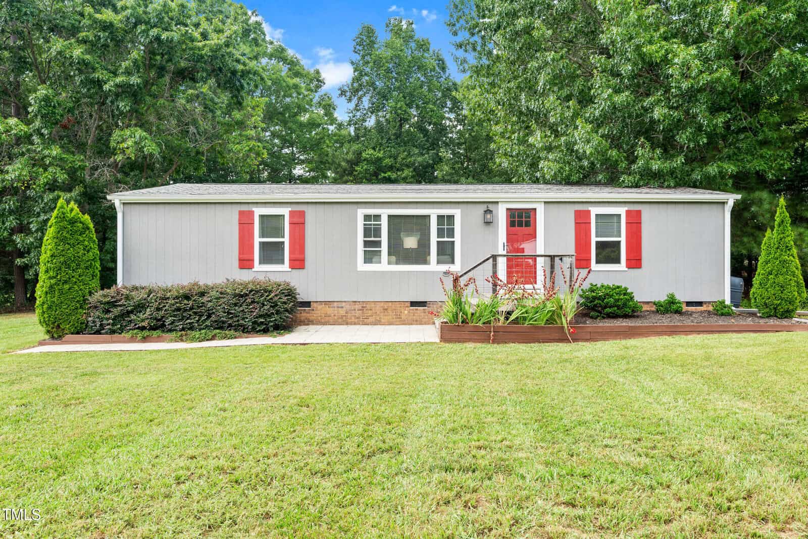 Single-story gray house with red shutters and a red front door, surrounded by green grass, bushes, and trees. A wooden ramp leads up to the entrance, and there are landscaped flower beds in front.