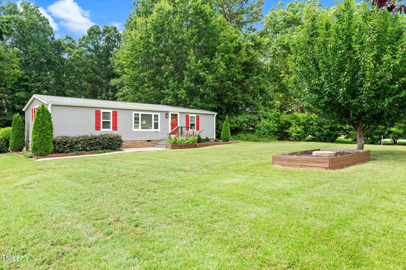 A single-story gray house with red shutters sits on a large, well-maintained lawn. There are green bushes near the house, a wooden raised garden bed, and trees in the background under a blue sky with clouds.
