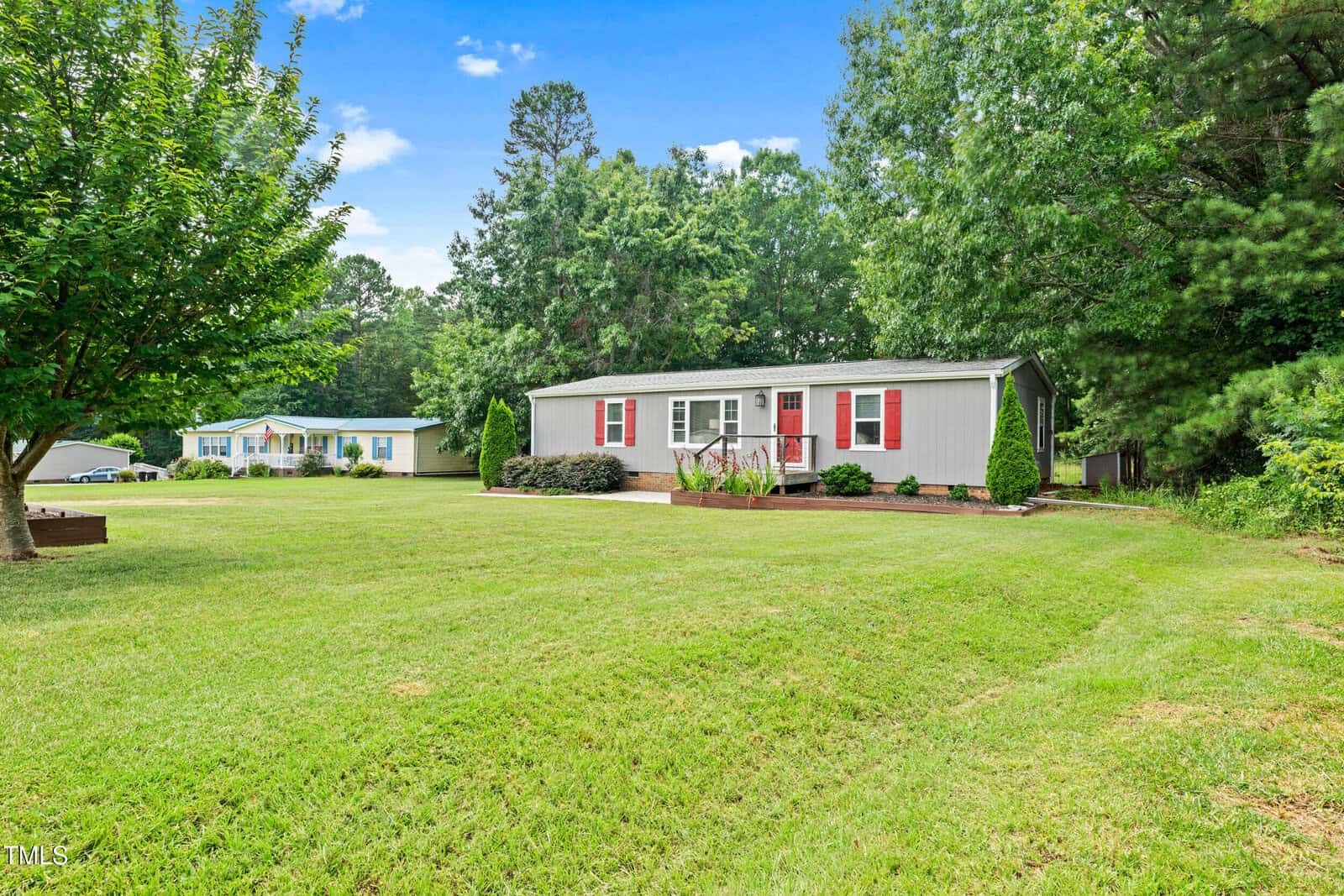 A single-story gray house with red shutters sits on a large grassy lawn, surrounded by trees and bushes, with another similar house visible in the background under a blue sky.