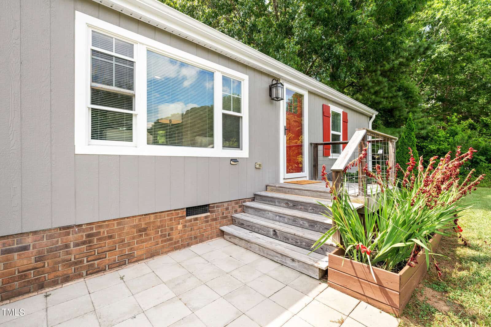 Small gray house with brick foundation, white-trimmed windows, and red shutters. Wooden steps lead up to the front door, next to a planter with tall red flowers. The patio is paved with square tiles and surrounded by greenery.