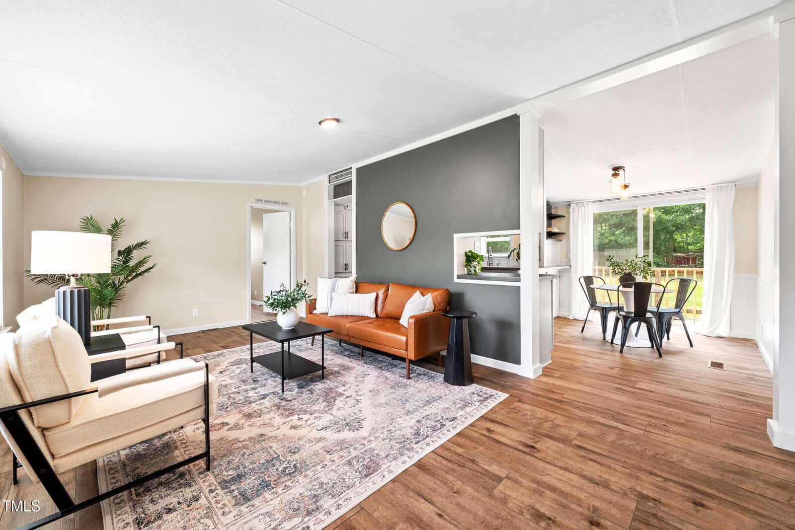 Modern living room with a brown leather sofa, two white armchairs, a patterned rug, and a black accent wall; dining area with a round table and four chairs is visible through an open doorway.