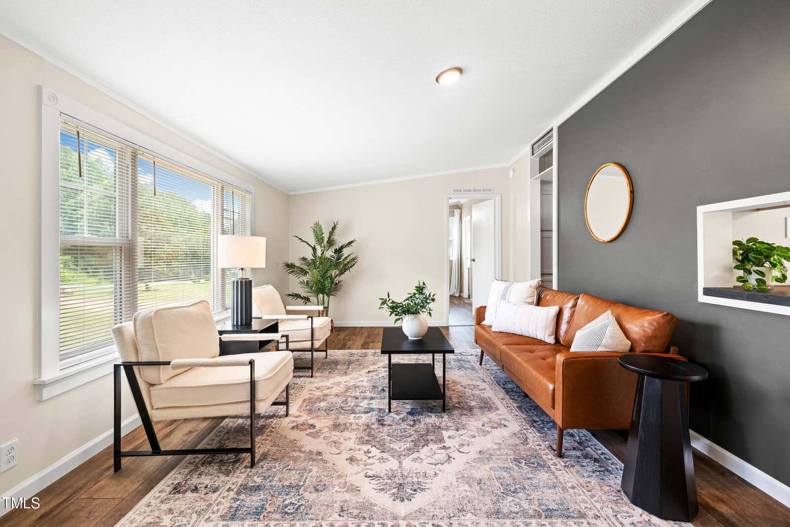 Modern living room with a brown leather sofa, two beige armchairs, a patterned area rug, coffee table, indoor plants, and large windows letting in natural light. One wall is dark gray with a round mirror and a built-in shelf.