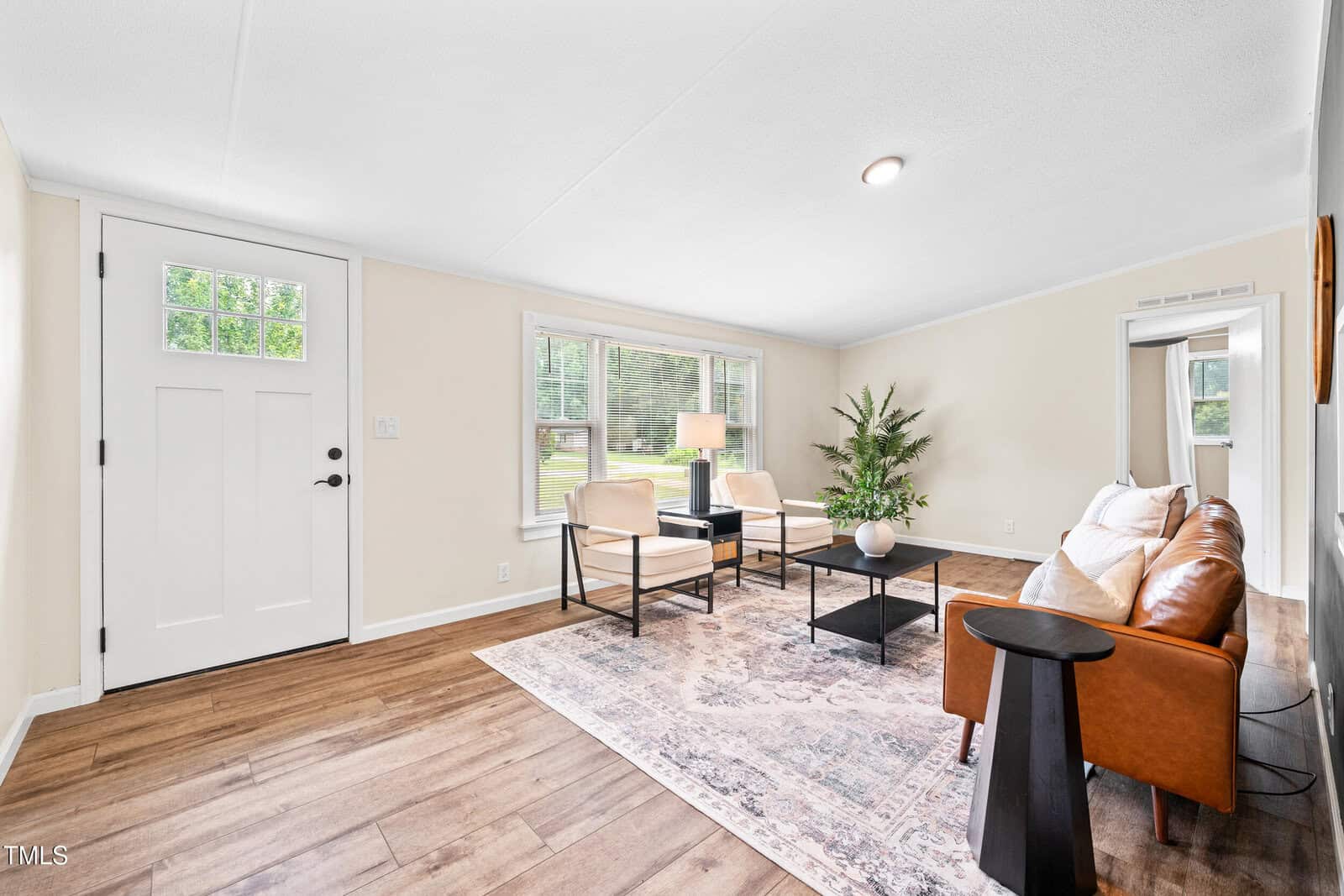 Bright living room with a brown leather sofa, two beige chairs, a patterned rug, a black coffee table, a potted plant, large windows, and a white front door. The space has wood flooring and neutral-colored walls.