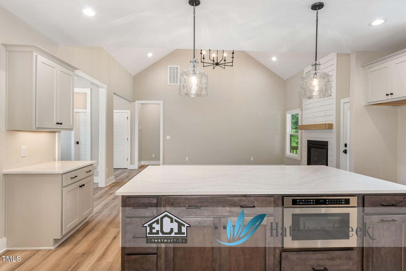 Modern kitchen interior with light beige walls, white cabinets, a large island with marble countertop, built-in appliances, pendant lights, wood flooring, and a view into an empty living area with a fireplace.