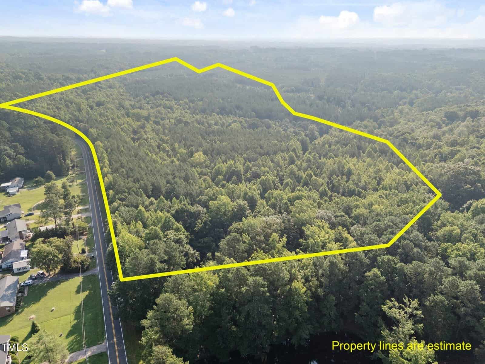 Aerial view of a large wooded area bordered by a yellow outline, indicating estimated property lines. A road and several houses are visible to the left of the outlined land. Text in the corner notes lines are an estimate.