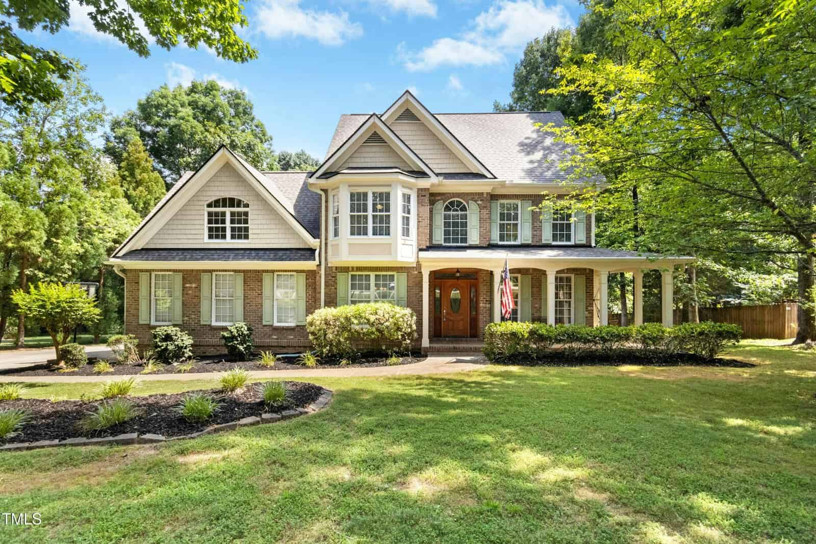 A two-story suburban house with beige siding, brick accents, and a large front porch. The home is surrounded by trees, with a well-maintained lawn and landscaped shrubs in the front yard.