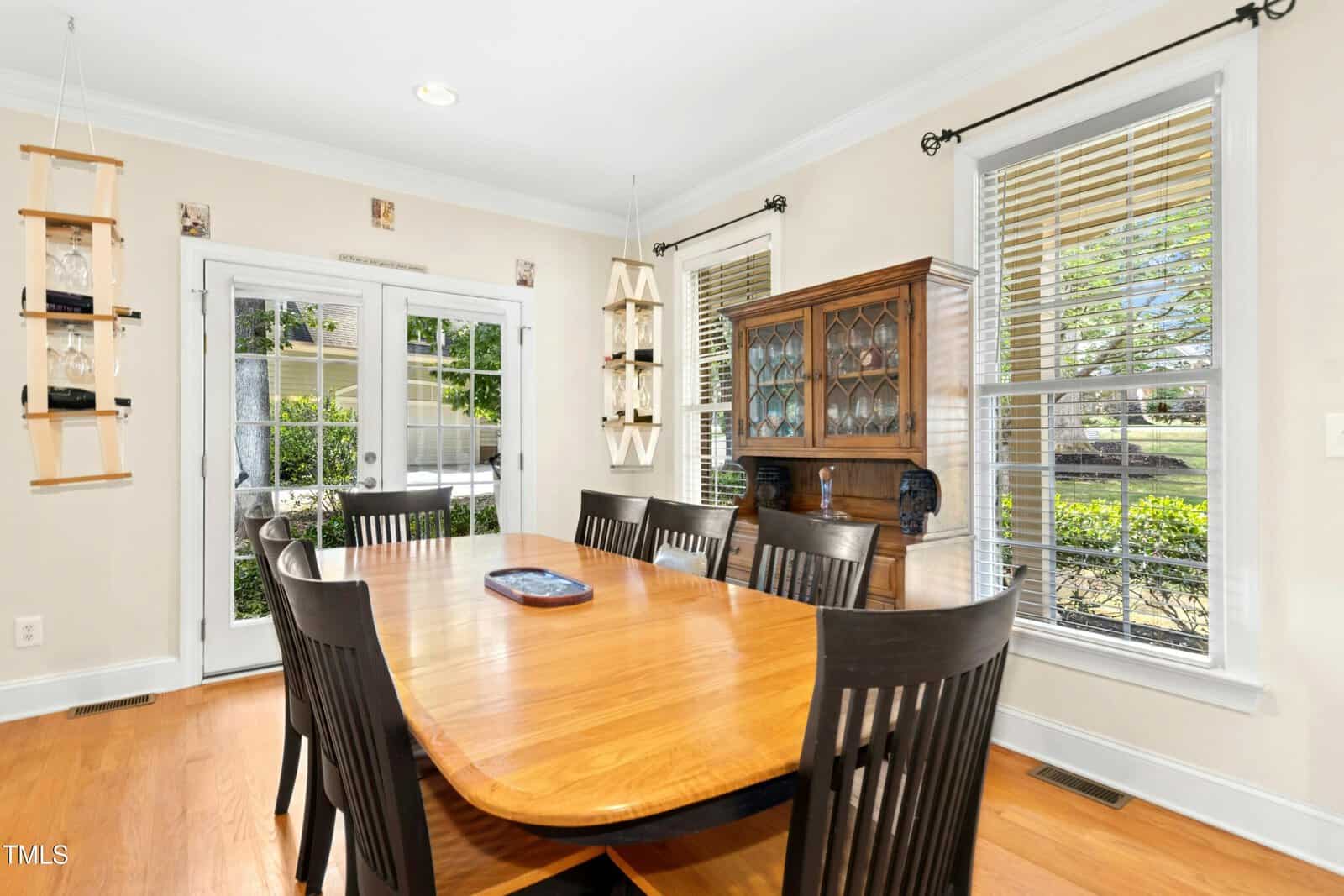 A bright dining room with a wooden table and eight dark chairs, a china cabinet, large windows with white blinds, and glass doors leading to a green backyard. Natural light fills the space.