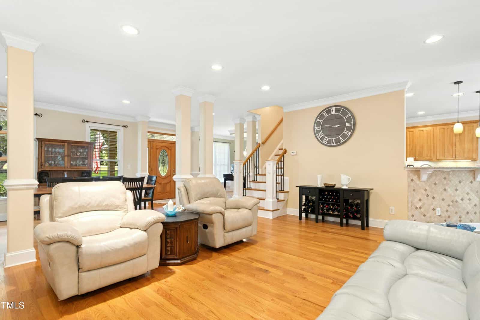 Bright living room with tan walls, hardwood floors, leather sofas, a wood coffee table, large wall clock, wine rack, and open view to dining area, wooden staircase, and kitchen with light-colored cabinets and tiled backsplash.