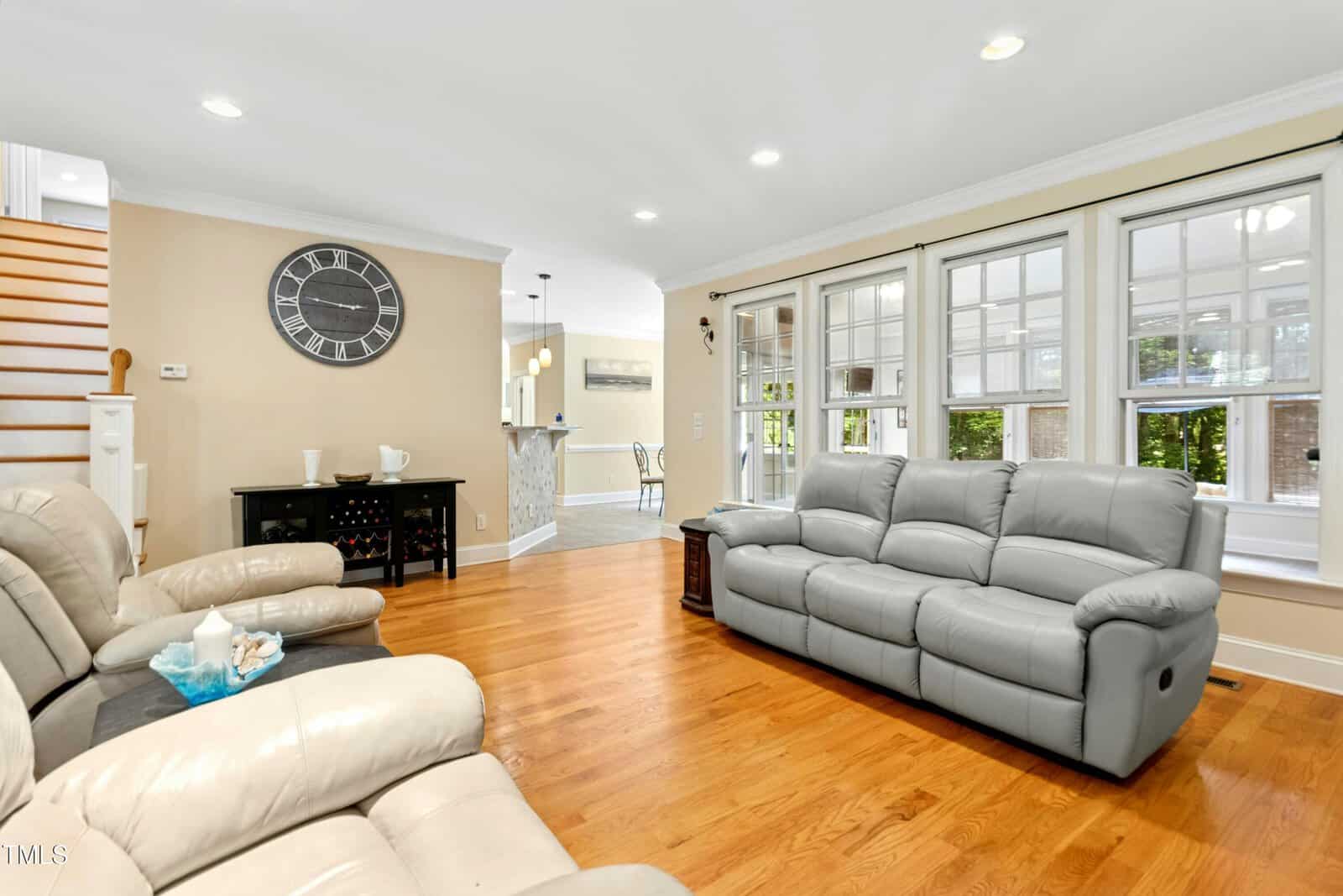 A bright living room with hardwood floors, two light gray leather sofas, a black sideboard with a round wall clock above, and large windows letting in natural light. Stairs and a kitchen area are visible in the background.