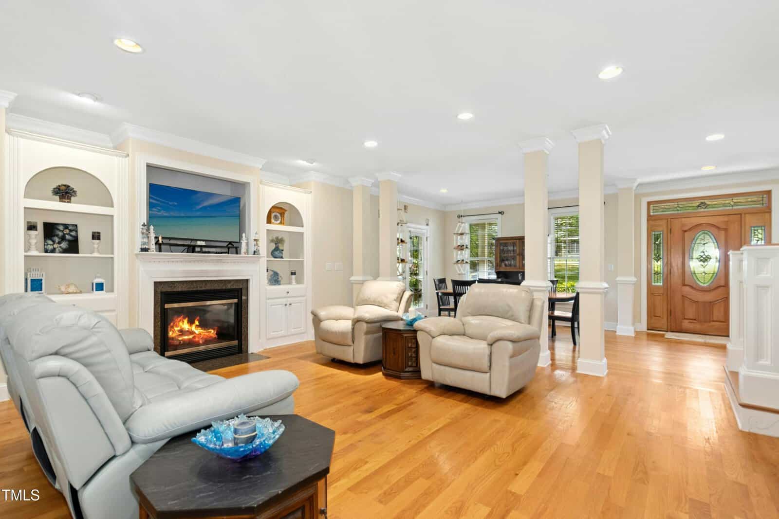 Bright living room with hardwood floors, a fireplace, two recliners, a sofa, built-in shelves, a flat-screen TV, round table, and an entryway with glass-paneled door. Large windows let in natural light.