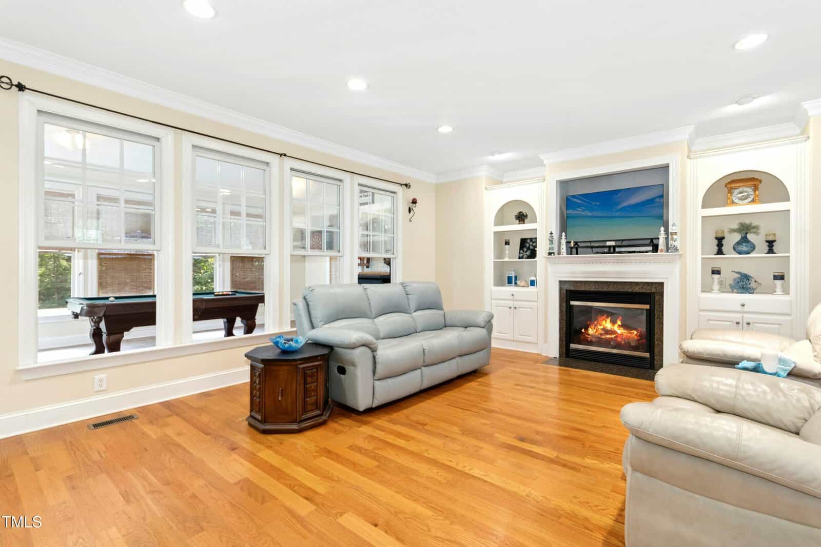 A bright living room with light wood floors, a gray leather sofa set, a lit fireplace, built-in shelves, and large windows showing a pool table in an adjacent sunroom.