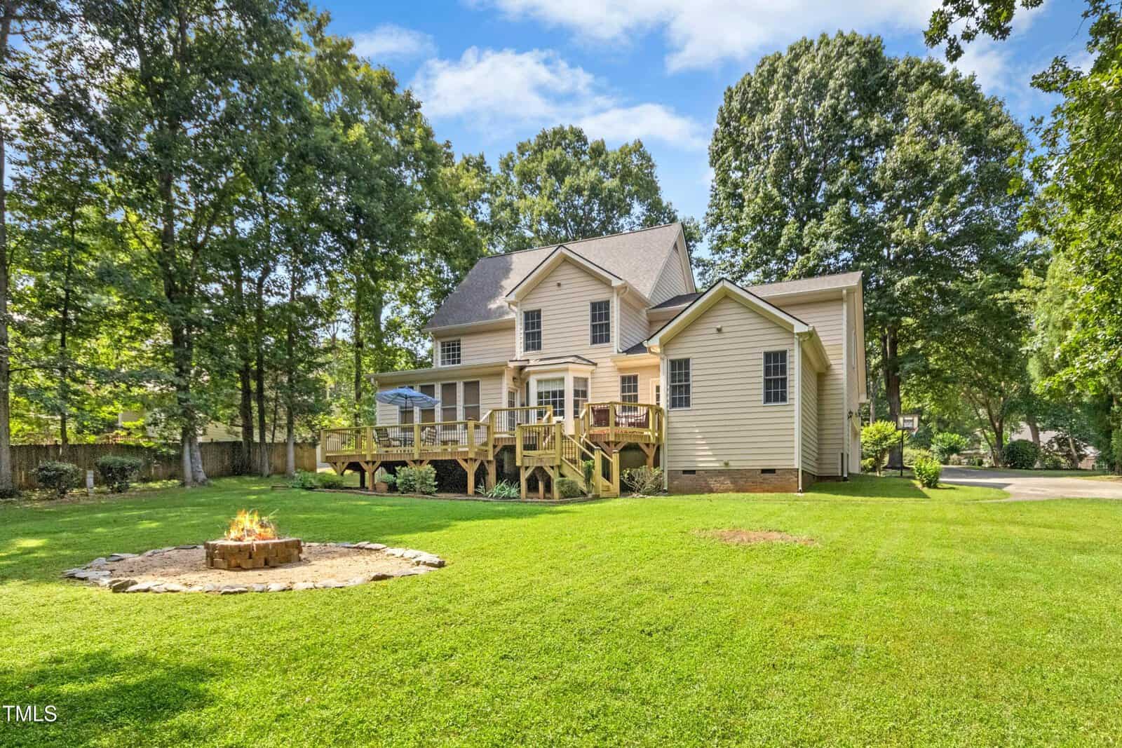 A two-story beige house with a wooden deck sits in a large, green backyard. A stone-ringed fire pit burns on the grass, surrounded by tall trees on a sunny day.
