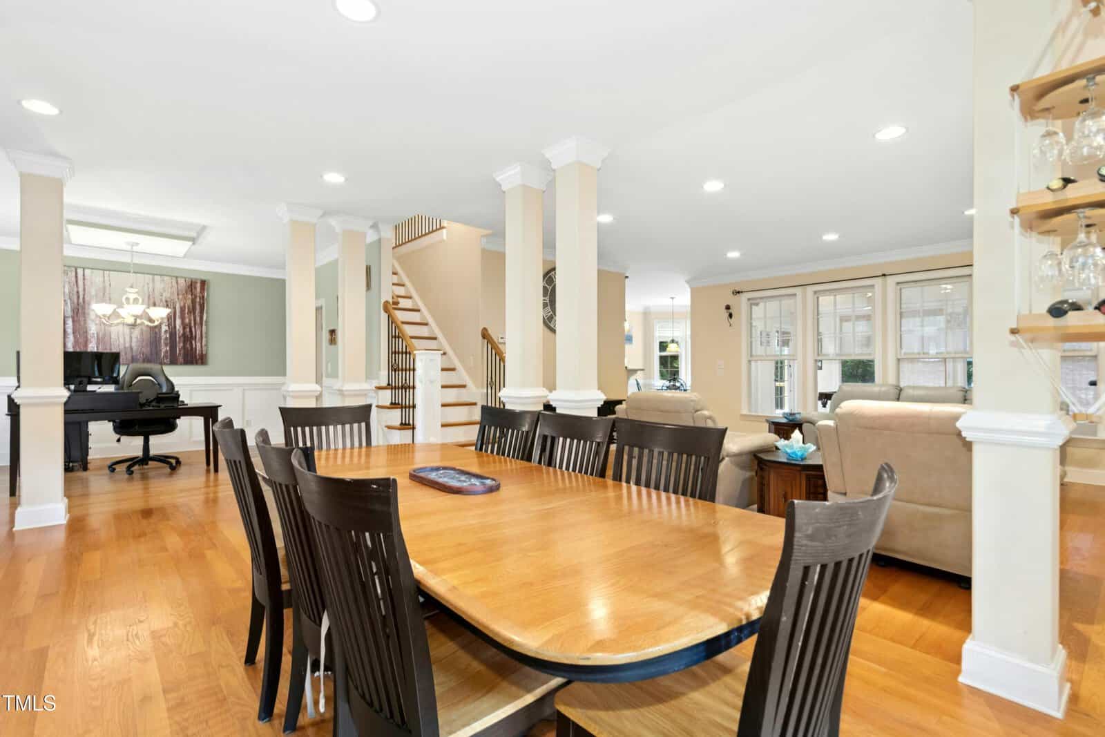 Open-concept living and dining area with a wooden table and dark chairs, beige sofas, hardwood floors, white columns, staircase, and a home office area with a desk in the background. Natural light fills the space.