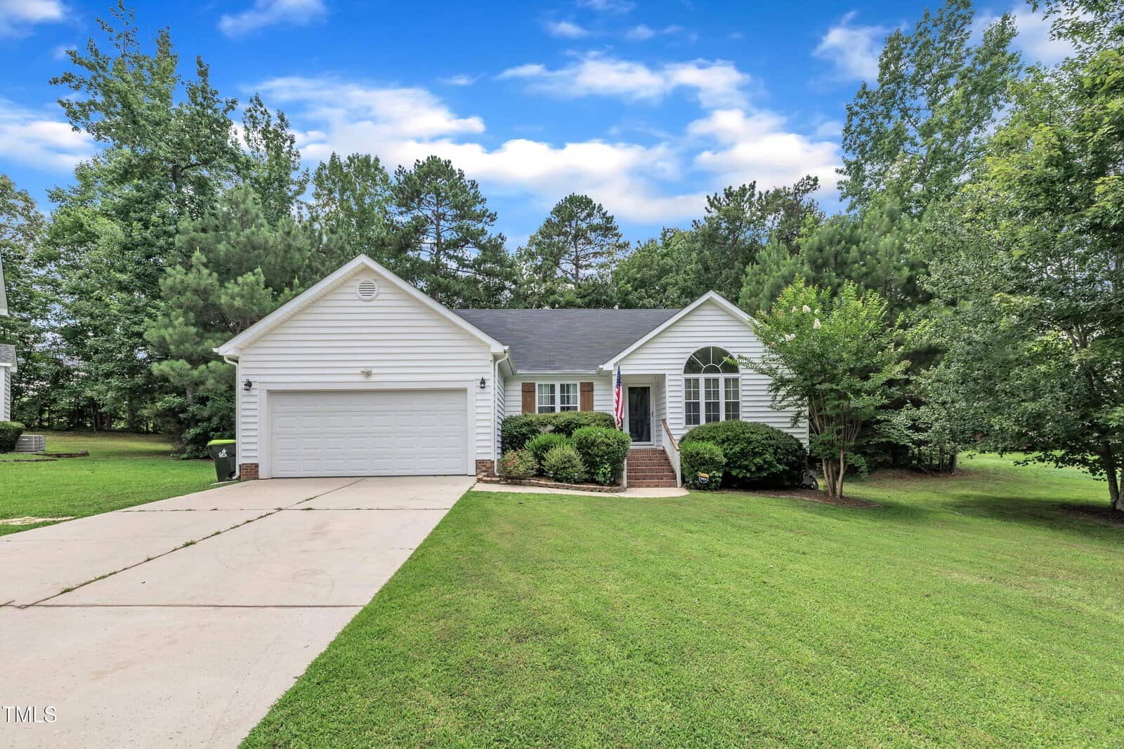 Charming single-story white house in Creedmoor at 2003 Zbonack Court, with a two-car garage, front steps leading to the entrance, neatly trimmed bushes, and a large grassy lawn surrounded by trees under a partly cloudy sky.