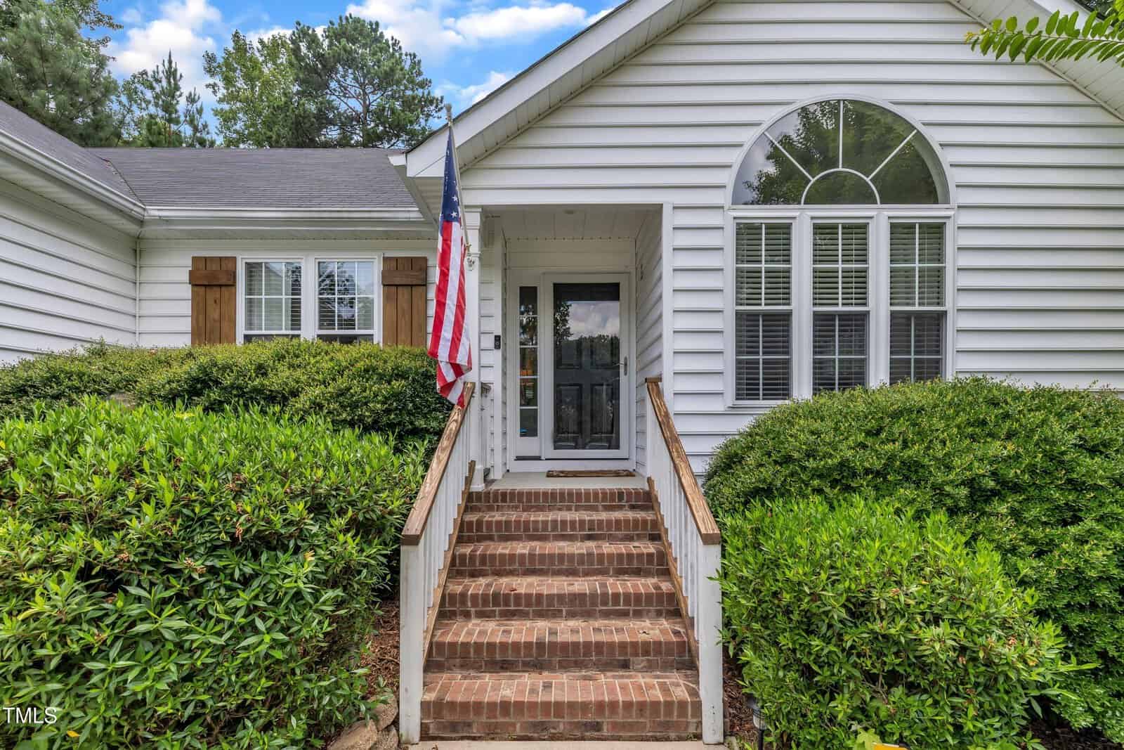 Front entrance of a white house with brick steps, white railing, and a glass storm door. An American flag is mounted by the door, and there are trimmed green bushes on both sides of the steps.