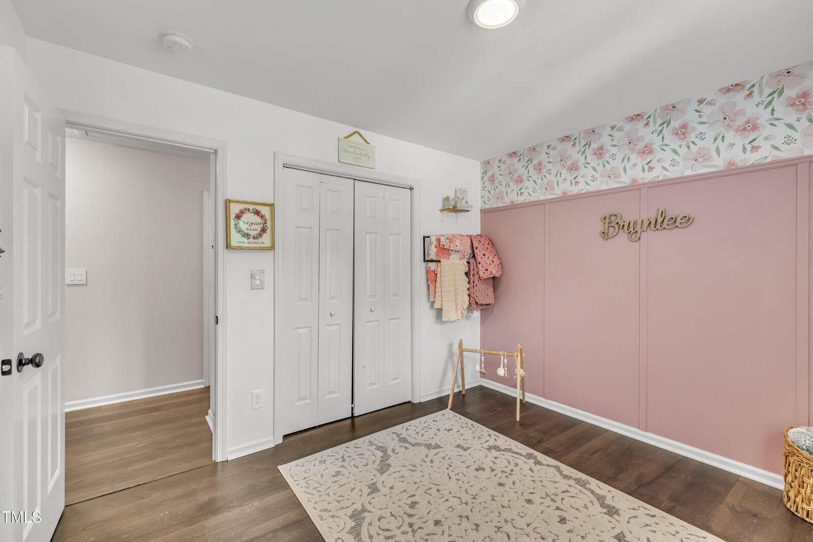 A children's room with floral wallpaper and pink paneling on one wall, a name sign reading "Brynlee," hanging clothes, a basket, a decorative rug, and double white closet doors.