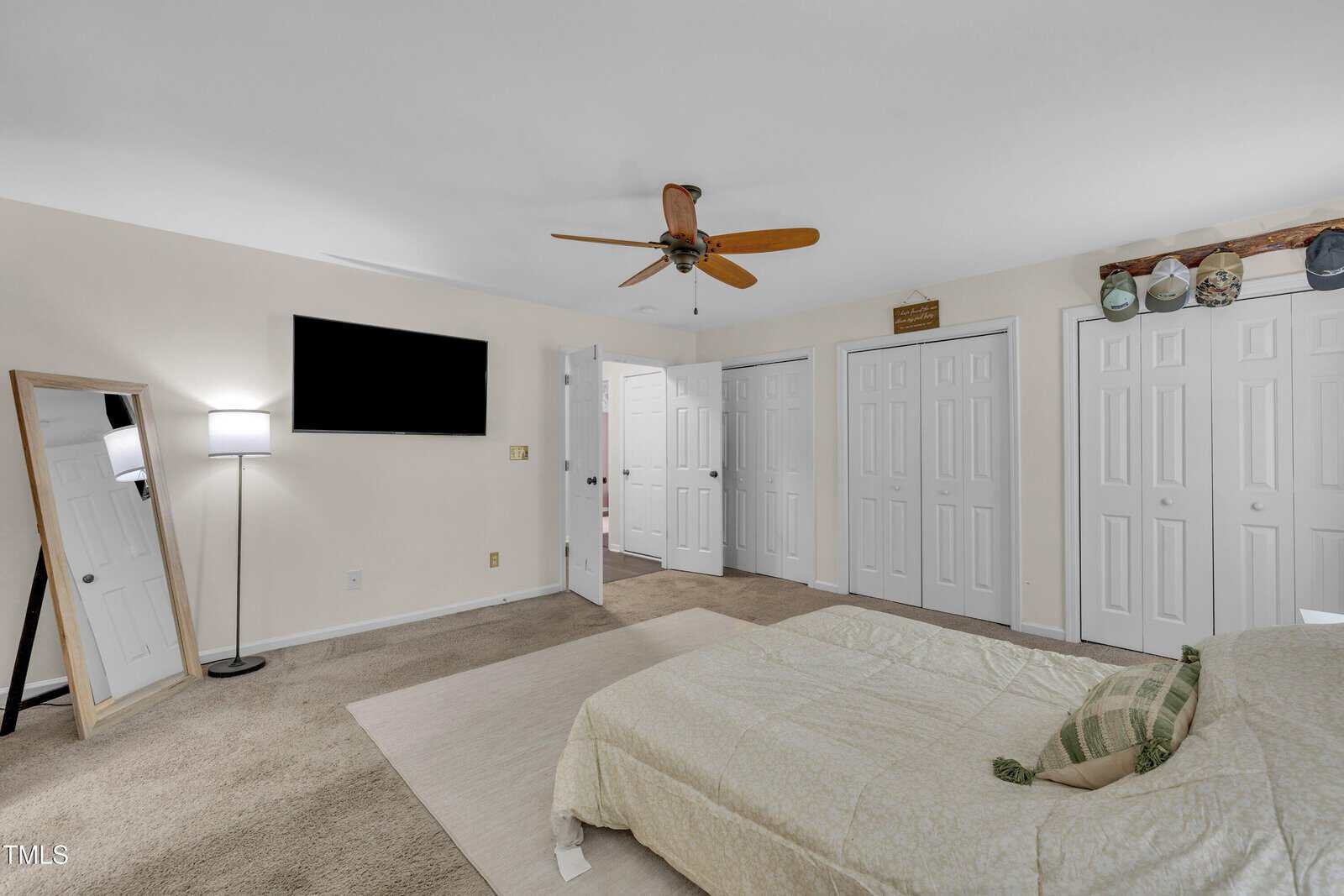 A bedroom with beige carpet, a bed with a light-colored comforter, a wall-mounted TV, a standing mirror, a floor lamp, multiple white closet doors, and a ceiling fan with wooden blades.