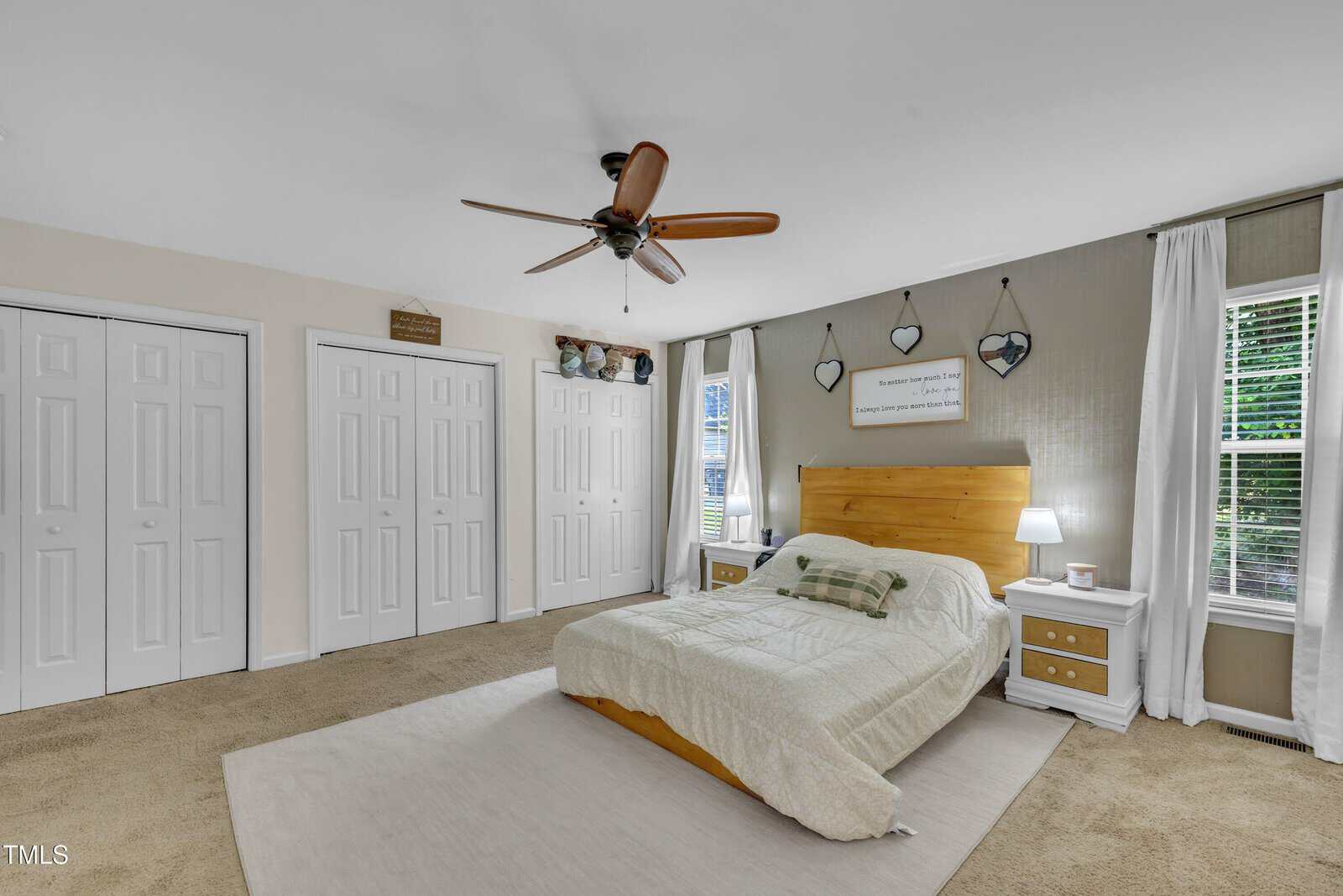 A cozy bedroom with a wooden bed, white bedding, two nightstands, a ceiling fan, three closets, and wall decor above the bed. Two windows let in natural light on either side of the bed.