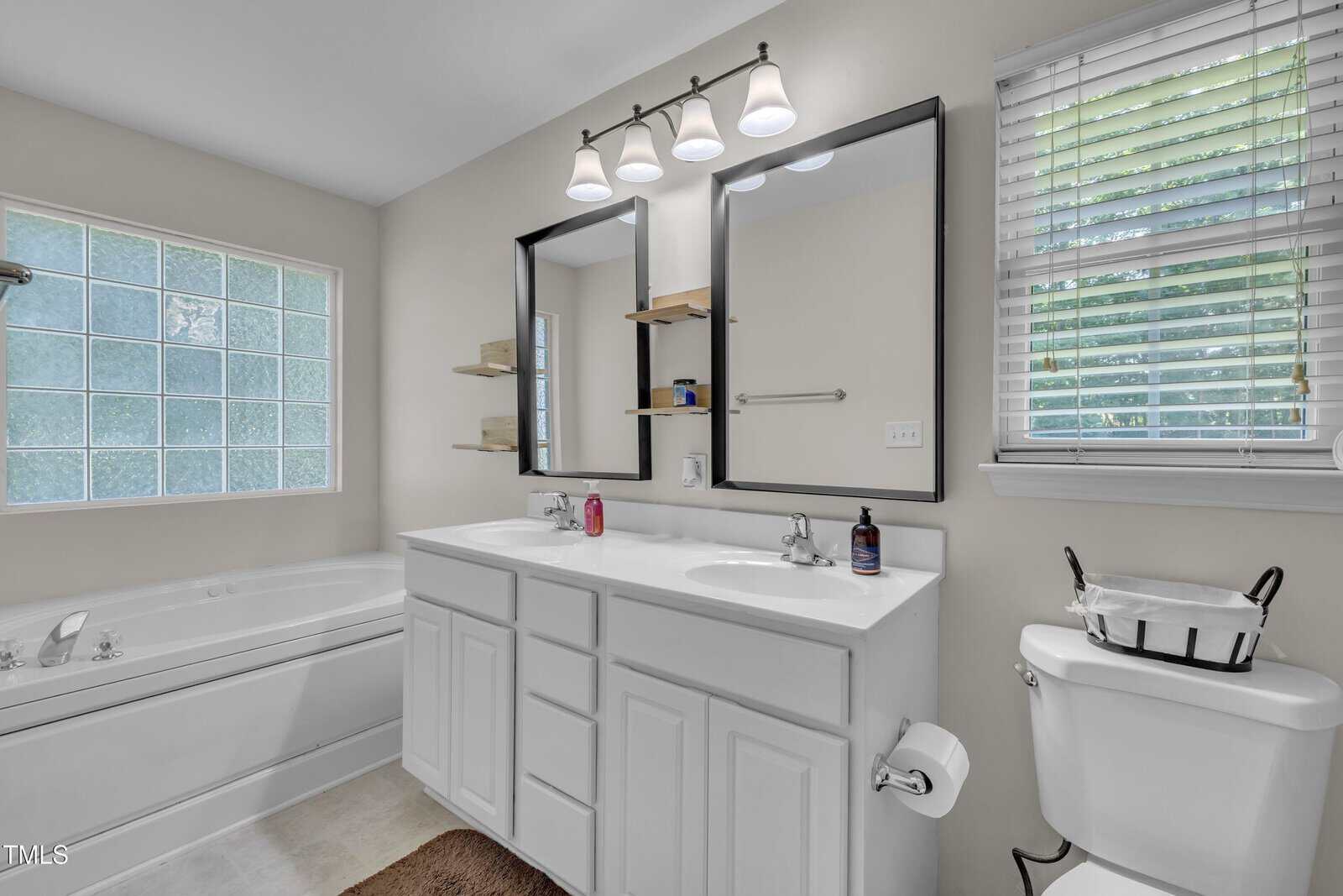 Modern bathroom with white cabinets, double sink, mirrors, bathtub, shelves, and a window with blinds letting in natural light.