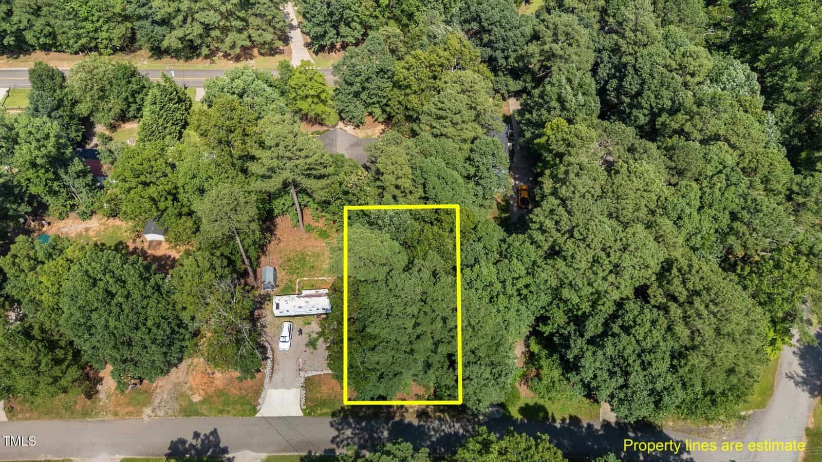 Aerial view of a wooded residential lot with a yellow box outlining part of the property. A small building and vehicle are visible near the bottom left. Streets border the lot, and trees surround the area. Text notes property lines are estimated.