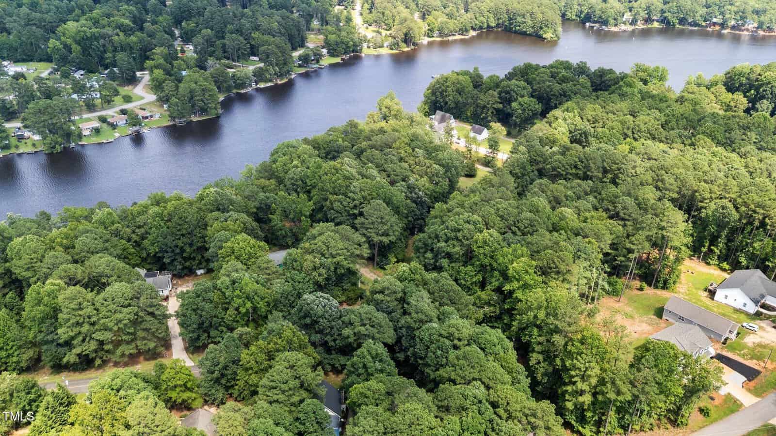 Aerial view of a wooded residential area near a lake, with houses scattered among dense trees and waterfront properties along the shoreline. Roads wind through the neighborhood and the surrounding landscape is lush and green.
