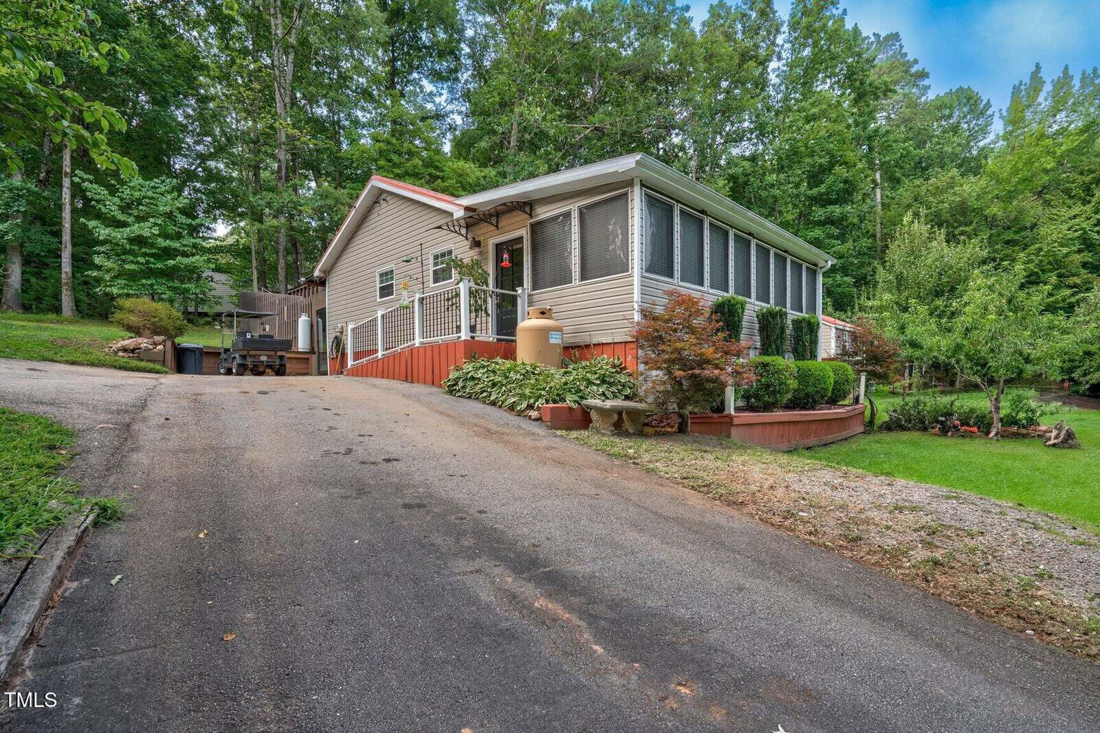 A light-colored manufactured home with a screened porch sits on a sloped driveway surrounded by lush green trees and landscaped bushes. The yard is well-maintained with a mix of grass, stone, and mulch beds.