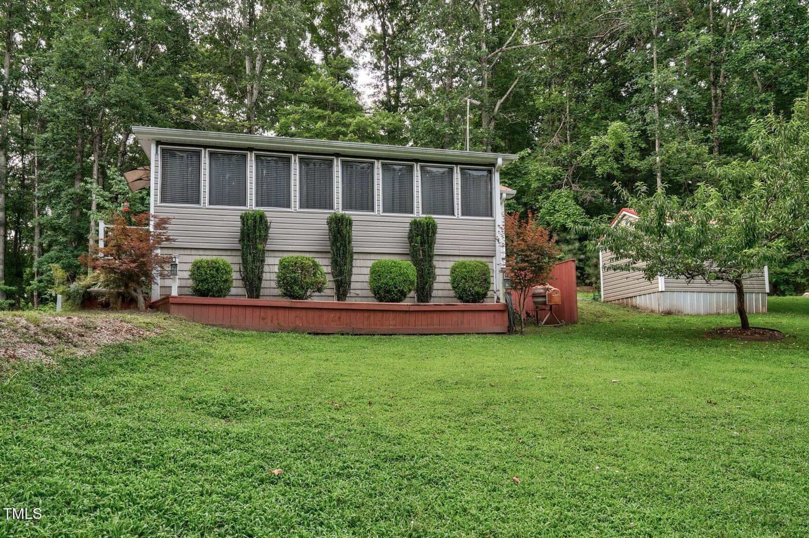 Single-story house with large windows and a raised wooden deck, surrounded by neatly trimmed bushes and trees, set on a grassy yard with a forested background.