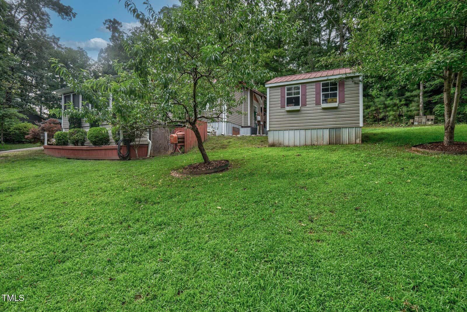 A green, grassy yard with a small tree, a light gray house with a porch on the left, and a small gray shed with pink shutters on the right. The area is surrounded by leafy trees under a partly cloudy sky.