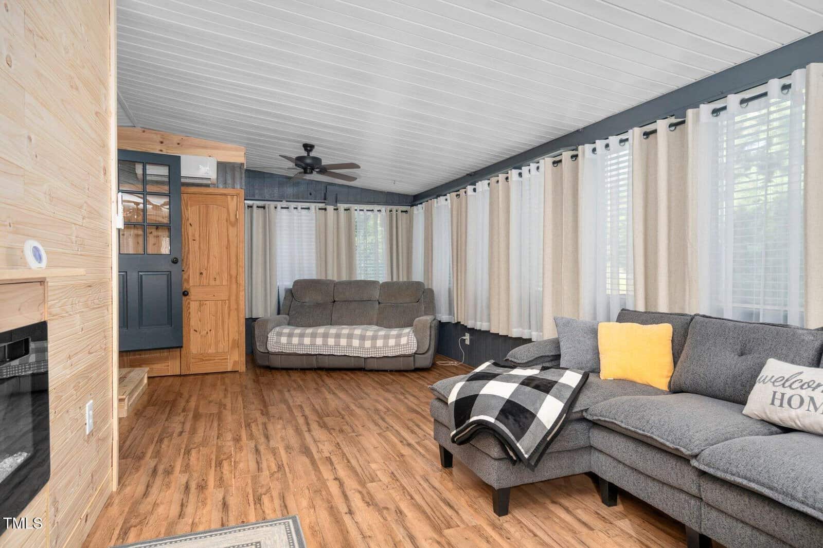 A cozy living room with wood floors, gray sectional sofa with pillows and a checked throw, a loveseat, large windows with white curtains, and a black ceiling fan. The walls are dark with light wood accents.
