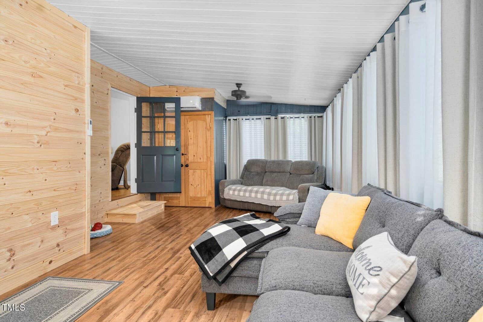 A cozy living room with wood-paneled walls, hardwood floors, a gray sectional sofa with pillows and a blanket, a futon in the background, and large windows covered with white curtains.