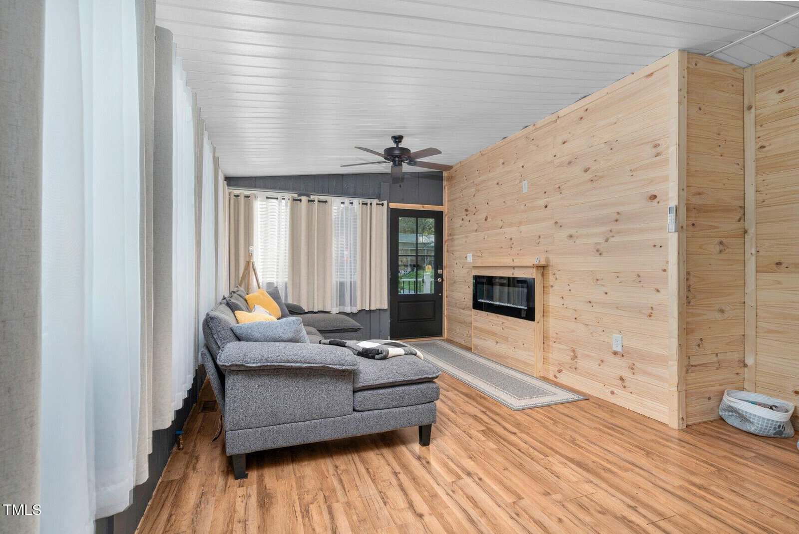 Modern living room with wood-paneled walls, a gray sectional sofa, light wood flooring, an electric fireplace, and a ceiling fan. Large windows with white curtains line one side of the room. A door leads outside.