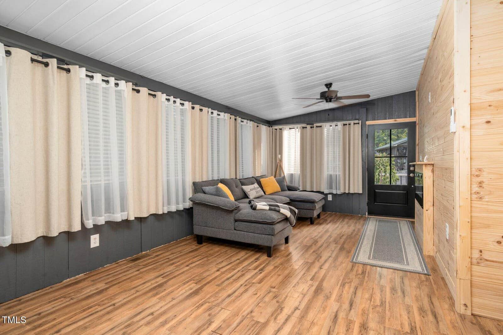 A living room with wood flooring, dark-painted walls, light curtains, a gray sectional sofa with pillows and throws, a ceiling fan, and a black door with window panels next to a wooden accent wall.