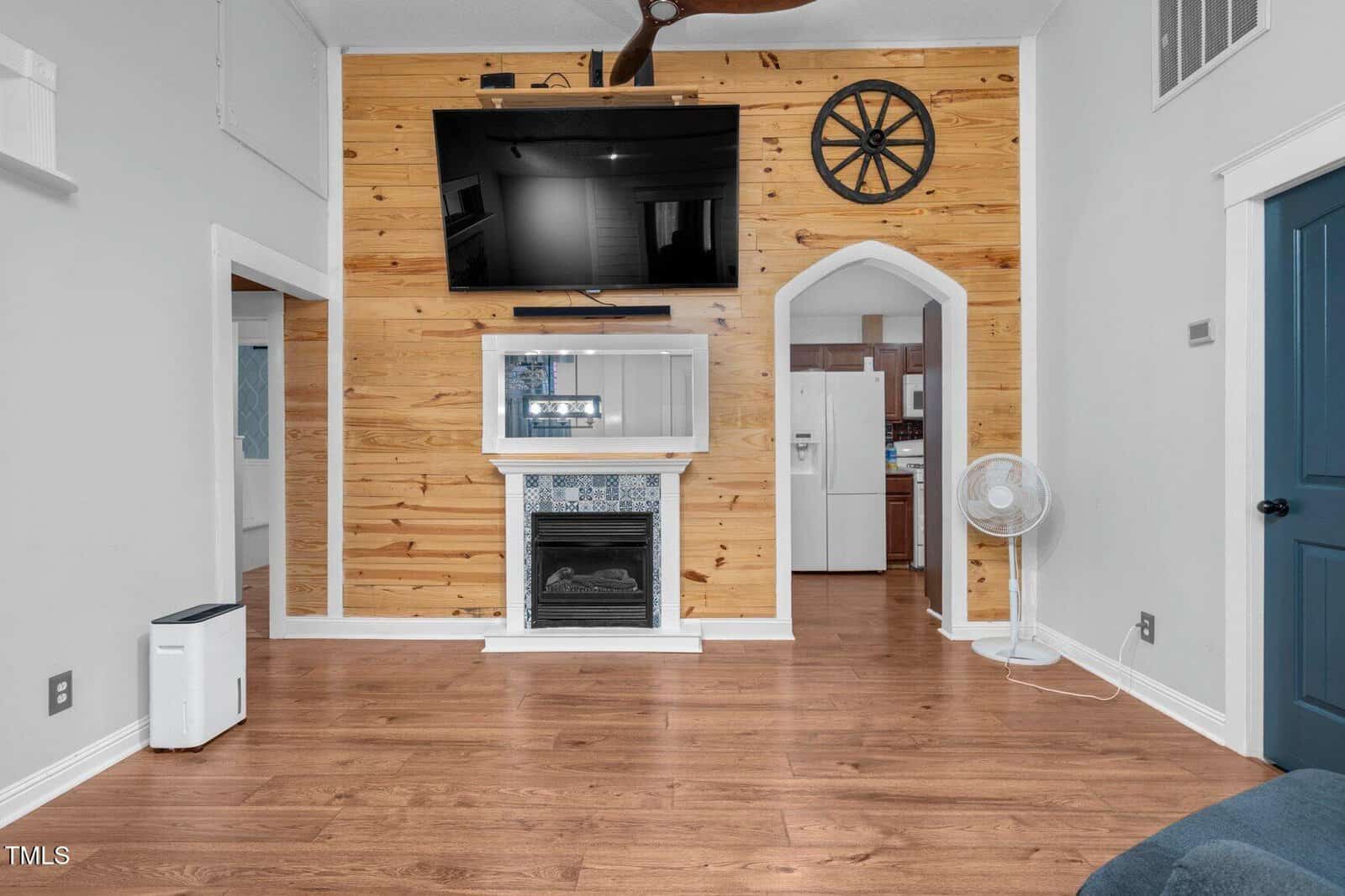 Living room with wood floor, a wooden accent wall featuring a mounted TV above a decorative fireplace, and an open doorway leading to a kitchen area with a fan and visible appliances.