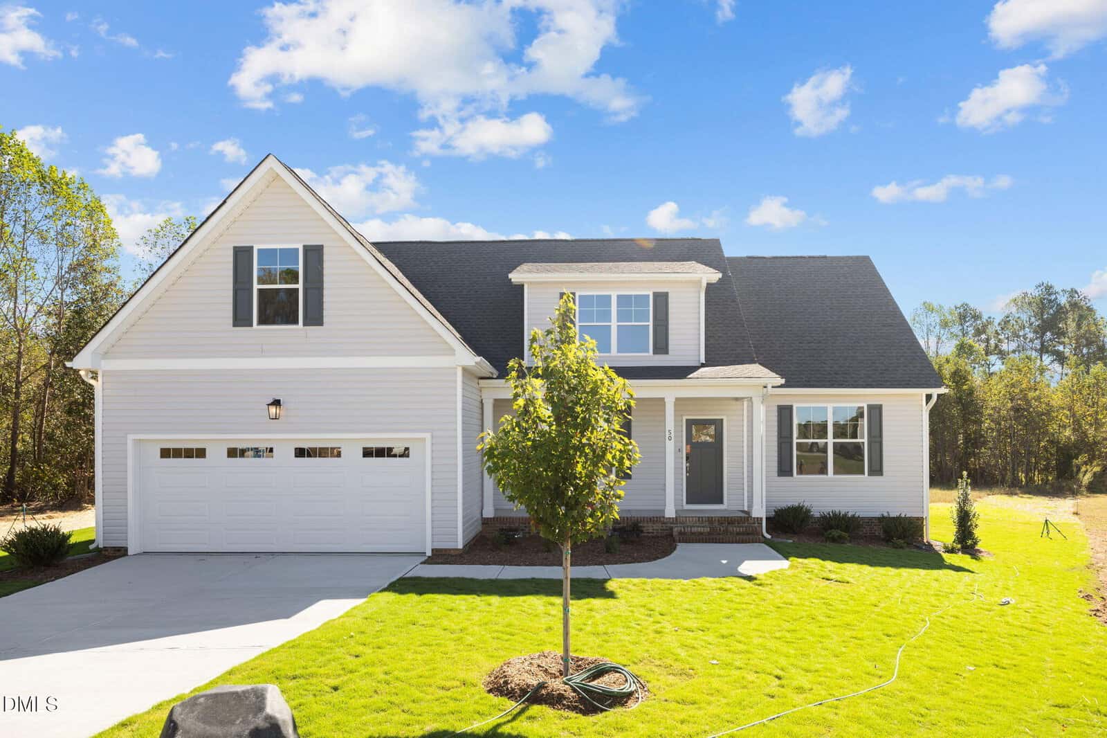 A modern, light-colored house with a two-car garage, front porch, and gabled roof sits on a neatly manicured green lawn under a blue sky with scattered clouds. A young tree is planted in the front yard.