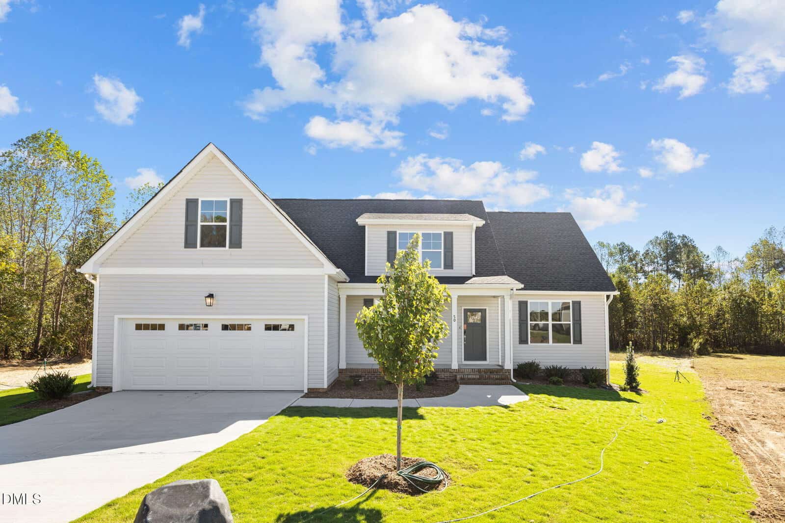 A modern two-story white house with black roof, double garage, front porch, and a small tree in a well-kept green lawn under a blue sky with scattered clouds.