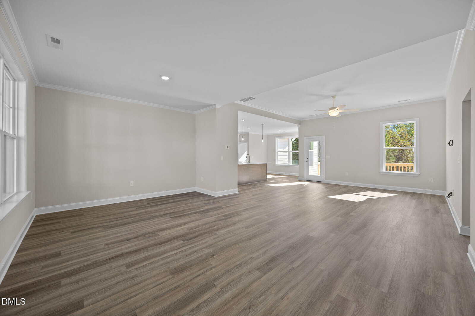 Spacious empty living area with light wood flooring, large windows, light gray walls, and open layout leading to a kitchen and patio door with outside view.