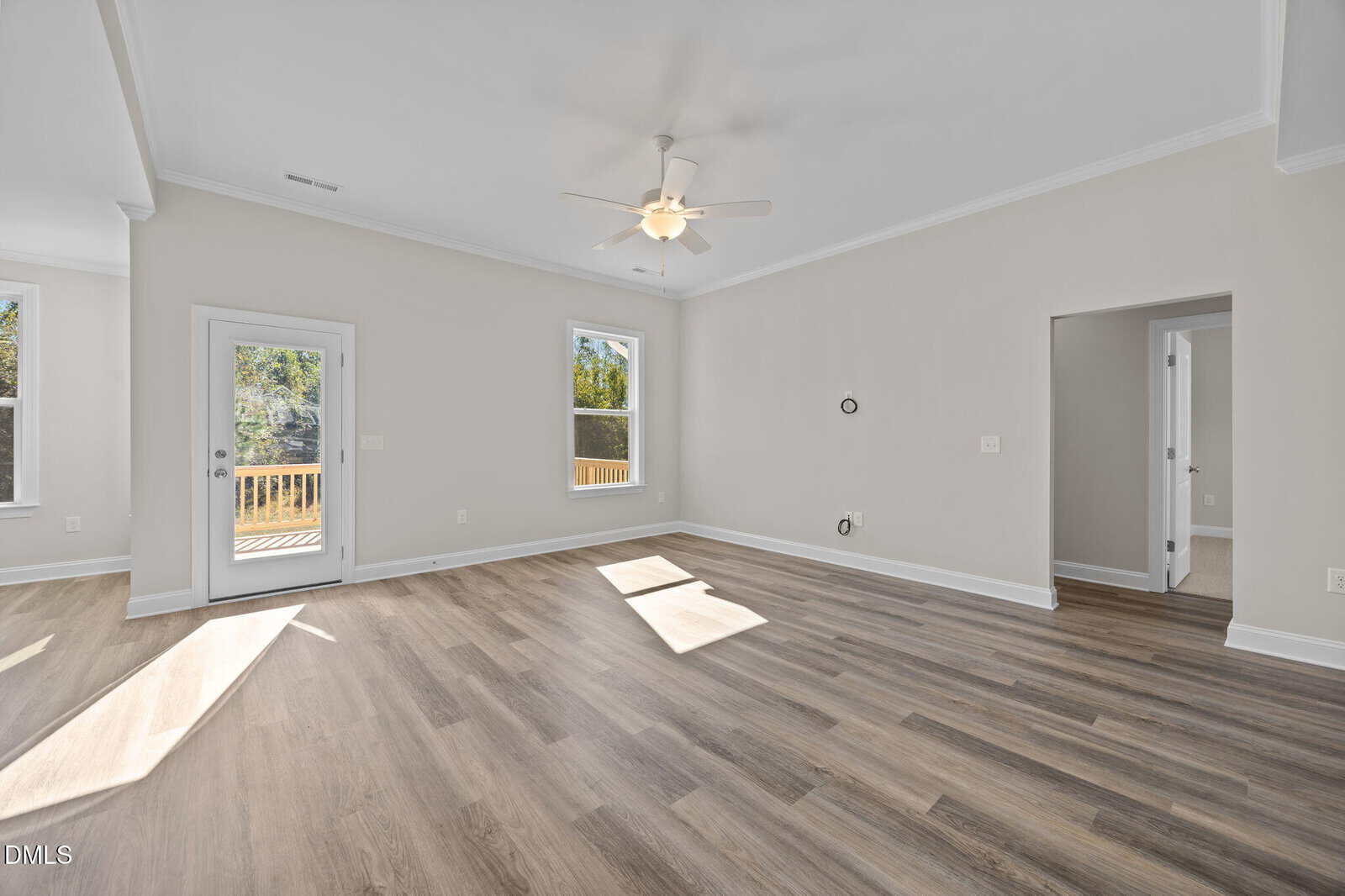 Bright, empty living room with light wood flooring, beige walls, ceiling fan, and two large windows. A glass door opens to a deck outside. An open doorway leads to another room.