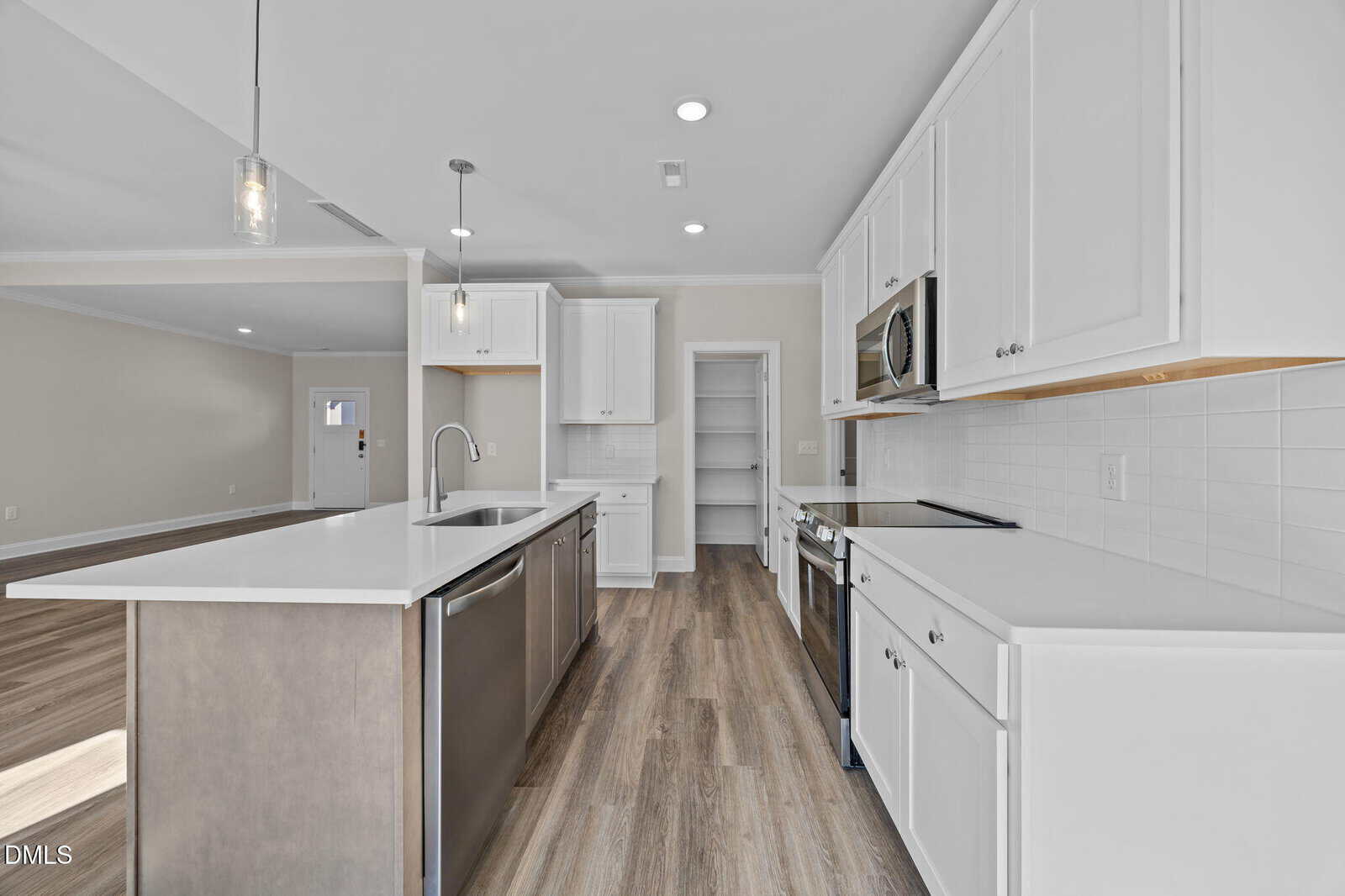 Modern kitchen with white cabinets, stainless steel appliances, a center island with sink, light wood flooring, recessed lighting, and an adjacent open living area. A pantry with built-in shelves is visible in the background.