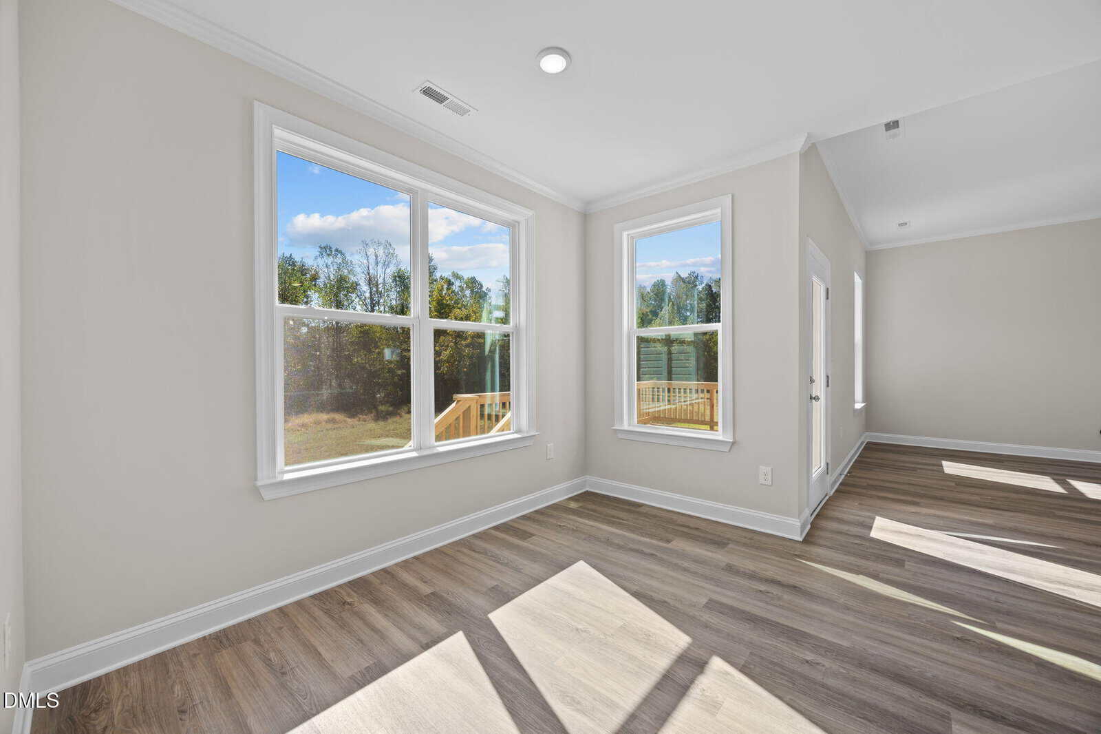 Bright room with large windows, light-colored walls, and wood flooring. Sunlight streams in, illuminating the space. Outdoor greenery is visible through the windows, and a glass door leads outside.